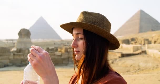 Frau mit Hut, wir löschen unseren Durst aus einer Plastikflasche, trinken gierig Wasser in der Wüste von Kairo, vor dem Hintergrund der Sphinx und der ägyptischen Pyramiden. Klarer, sonniger Tag, Hitze, Zeitlupe — Stockvideo