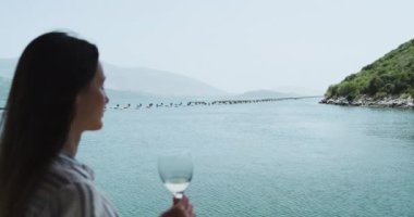 Uzun saçlı güzel bir kız, bir deniz midyesi çiftliğinin arka planında beyaz şarap içiyor. Deniz çiftliğine odaklan, kız bulanık. Açık gün, yavaş çekim, orta çekim