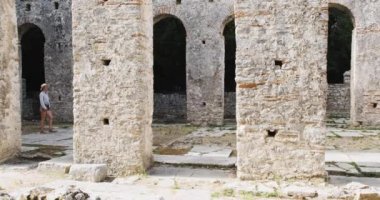 Kız, antik şehir Butrint 'in kalıntılarının yanından geçiyor, binaların taş sütunlarının yanından geçiyor. Temiz gün, geniş açı. Yüksek kalite 4k görüntü