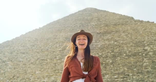 Jonge vrouw in een hoed dansend voor de piramide van Gizeh in Caïro, gelukkig, glimlachend. Medium schot, slow motion, heldere zonnige dag — Stockvideo