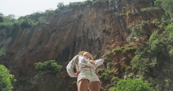 Young girl in a hat and denim shorts and a shirt stands and looks up fearfully up in a gorge with green trees. Take your breath away in Albania at Gjipe Canyon. Medium shot, clear day, slow motion — Stock Video
