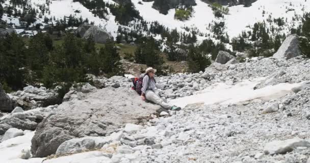 Young girl in a hat sat down to rest with a backpack on a rocky mountain path in the Alps. Against the backdrop of snowy mountains, green trees. Daytime, wide shot, slow motion — Stock Video