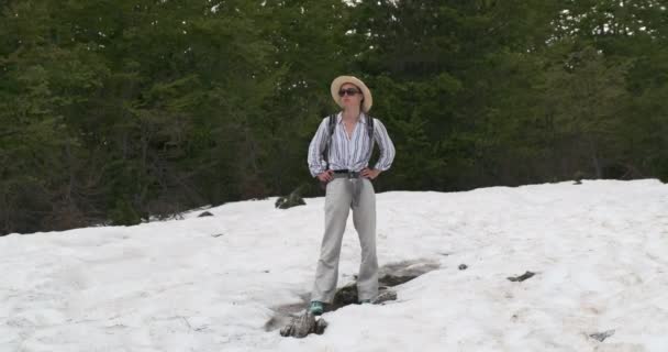 Νεαρή κοπέλα με καπέλο, γυαλιά ηλίου, πουκάμισο και παντελόνι, με σακίδιο, στέκεται στη μέση ενός πράσινου αλπικού δάσους, στο χιόνι και κοιτάζει γύρω της. Μέρα, άνοιξη, μέτρια βολή, Άλπεις — Αρχείο Βίντεο