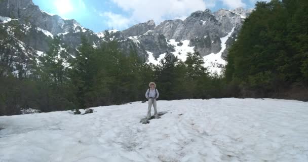 A young girl in a hat, sunglasses, a light shirt and trousers, with a backpack, stands among the green alpine forest on a snowy meadow and looks around. Against the backdrop of mountains, sun, blue — Stock Video