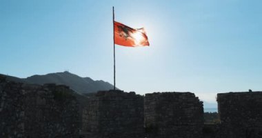 Kırmızı ve siyah Arnavut bayrağı eski kalenin üzerinde dalgalanıyor. İçinden parlak bir güneş doğuyor. Bağımsızlığın ve vatanseverliğin sembolü..