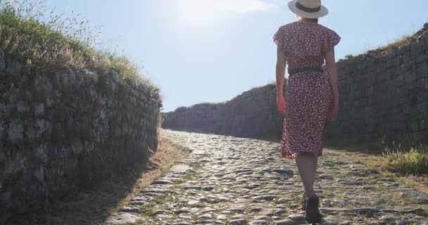 Schöne Mädchen in einem Kleid und einem Hut geht entlang einer alten Kopfsteinpflasterstraße in Richtung Sonne, Rückansicht, tagsüber, klarer Tag, untergehende Sonne. Schöner Wanderweg mit magischem Sonnenlicht — Stockvideo