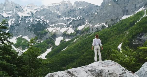 Krásná dívka, stojí na kopci, zvedá paže k bokům a objímá svět. Na pozadí albánských Alp se sněhem, zeleným lesem, širokými záběry, ve dne. Kouzelný pohled na hory — Stock video