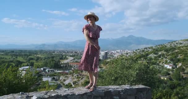 Beautiful Caucasian girl in a dress, hat stands on top, turns her back to the camera and spreads her arms to the sides, hugging the world. A magical view from the top of the green valley and mountains — Stock Video