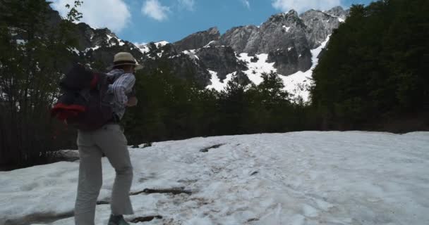 Tourist girl with a backpack climbs a path covered with snow in the Albanian Alps. Green forest and snow-capped mountains in the background. Daytime, blue sky, wide shot, spring in the mountains. — Stock Video