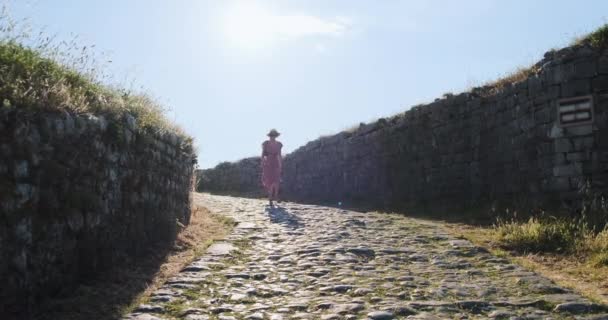 Piękna biała dziewczyna w sukience, kapeluszu i okularach przeciwsłonecznych, schodzi starożytną drogą kamieni, widok z przodu, dzień, jasny dzień, zachodzące słońce od tyłu. Kamienny ogrodzenie wzdłuż drogi — Wideo stockowe