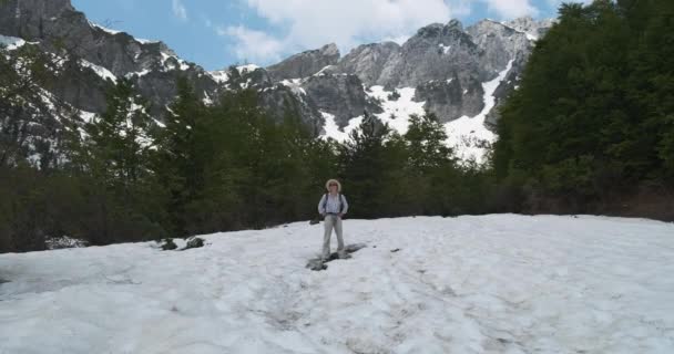 Una esbelta mujer vestida con ropa ligera, una turista con una mochila, se encuentra entre el verde bosque alpino en un prado cubierto de nieve y mira a su alrededor. Sobre el telón de fondo de las hermosas montañas de Albania, azul — Vídeo de stock