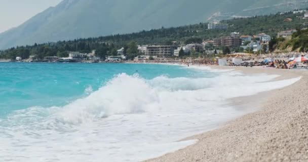 Miasto nadmorskie w Albanii. Ruch piankowych fal na żwirowej plaży. W oddali widać osoby z parasolkami plażowymi. Czyste błękitne niebo, szeroki strzał — Wideo stockowe