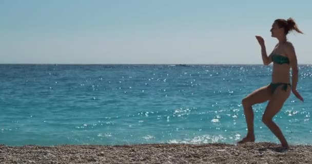 Piękna kaukaska dziewczyna w bikini bawi się samotnie na plaży, na tle idealnego turkusowego morza. Tańcząc jak Egipcjanin, z ramionami na boki. Dzień, szeroki strzał. — Wideo stockowe
