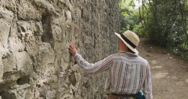 Jonge vrouwelijke toerist verkent de oude ruïnes van de stad Butrint in Albanië. Hij passeert een stenen muur en loopt met zijn hand over de stenen. Overdag, wijd schieten, boomschaduw — Stockvideo