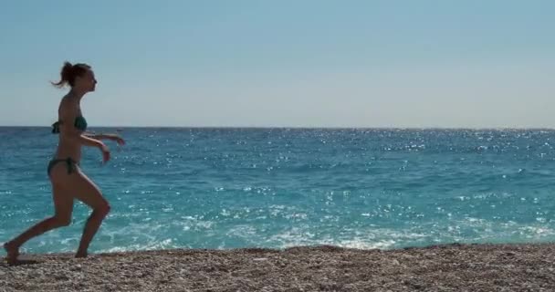 Junges schönes Mädchen tanzt am Meer entlang. Seitenansicht, klarer Tag, blaues Meer und Himmel, Kieselstrand, mittlerer Schuss — Stockvideo