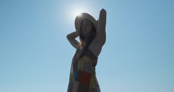 Młoda kobieta w sukience i kapeluszu, stojąca na wietrze przed słońcem, odwraca się, by się rozejrzeć. Trzyma się za ręce. Dzień, jasne słońce, błękitne bezchmurne niebo — Wideo stockowe