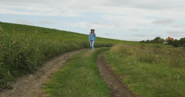 Жінка в джинсах і капелюсі йде по ґрунтовій доріжці вздовж поля з зеленою високою травою до камери. Хмарний день, повільний рух, широкий постріл — стокове відео