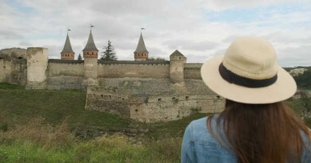 Πίσω όψη, γυναίκα με μακριά μαλλιά σε ένα καπέλο, κοιτάζει το αρχαίο κάστρο φρούριο. Μέρα, μέτρια βολή, συννεφιά. Kamenets Podolsky, Ουκρανία. — Αρχείο Βίντεο