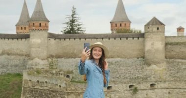 Şapkalı genç bir kız kuleleri olan antik bir kalenin arka planında telefonla selfie çekiyor. Orta çekim, gündüz, bulutlu, yavaş çekim