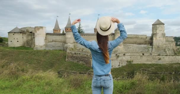 Young Girl se para frente a una antigua fortaleza-castillo, se quita el sombrero y extiende los brazos hacia los lados, abrazando al mundo. Kamenets Podolsky, Ucrania. Diurno, nublado, plano medio. — Vídeo de stock
