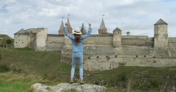 Achteraanzicht van een jonge vrouw in denim springend op een rots voor een oud fort. Het meisje zwaait met haar handen en heeft plezier met een prachtig uitzicht op het kasteel. Overdag, wijd open schot. — Stockvideo