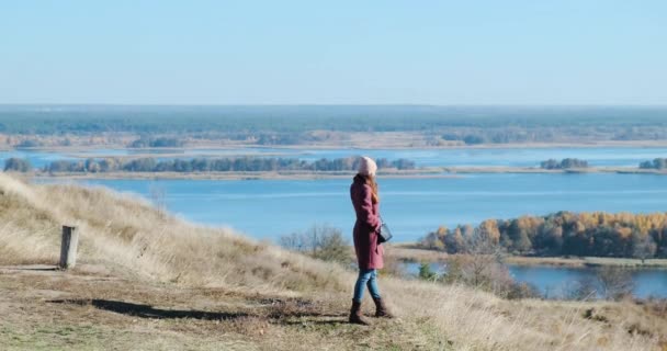 Girl in a marsala coat stands on the top of a plateau in autumn and talks on the phone against the backdrop of a wide river. Daytime, wide shot — Wideo stockowe