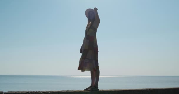 Young woman in a hat and sundress stands in full growth against the sun against the backdrop of the sea surface of the water. The dress develops in the wind. — Stok Video