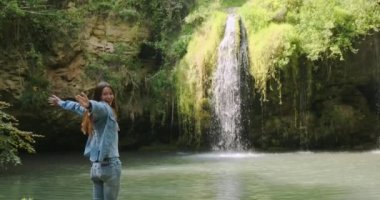 Uzun saçlı beyaz kız küçük bir şelaleyle bir lagünün önünde duruyor ve seviniyor. Ellerini sallıyor, orta plan, gündüz, yaz.
