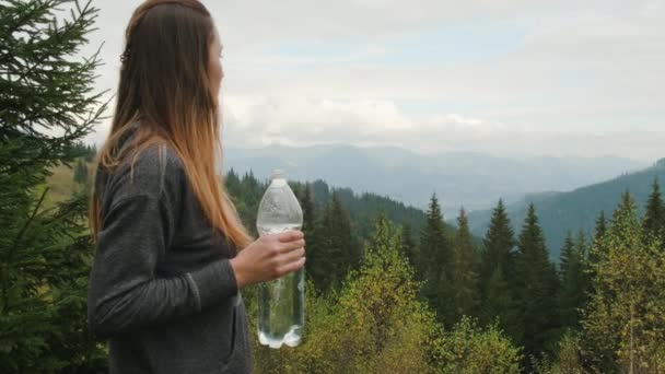 Caucasian girl, in the forest against the backdrop of mountains, drinks water from a plastic bottle. Green coniferous trees, medium plan, cloudy, slow motion — стоковое видео
