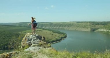 girl is on the edge of the cliff, spreading her arms to the sides, view from the back. Wide river, canyon, greenery, blue sky. Wide world to travel