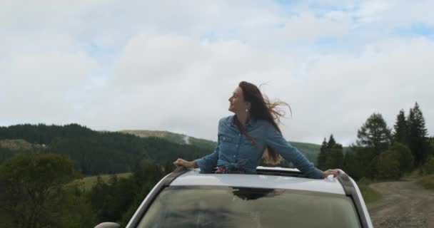 Bela menina de cabelos compridos inclinou-se para fora da escotilha de um carro estacionado nas montanhas. Olha em volta da floresta verde e da natureza. Tiro médio, geralmente — Vídeo de Stock