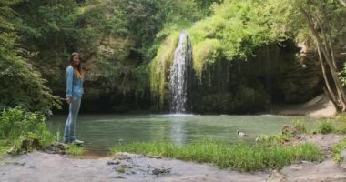 Kadın şelaleli küçük bir gölü işaret ediyor. Gündüz, geniş açı. Güneş şelalede parlıyor.