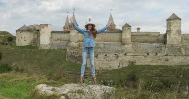Девушка прыгает на камне на фоне старого замка с башнями. Он машет руками, радуется. День, облака, Каменец Подольский, Украина — стоковое видео