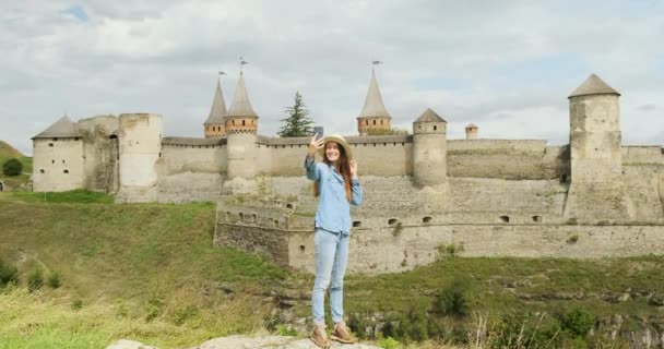 Девушка стоит в полном росте на камне на фоне древней средневековой крепости и фотографирует себя по телефону. — стоковое видео