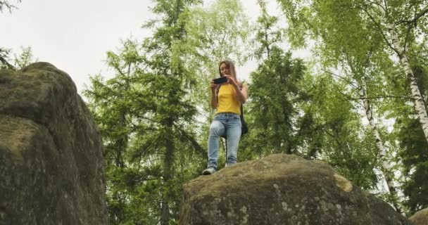 Young girl climbed a hill and takes pictures in the forest on her smartphone, standing on a stone. Green trees on the background, bottom view — Vídeo de Stock