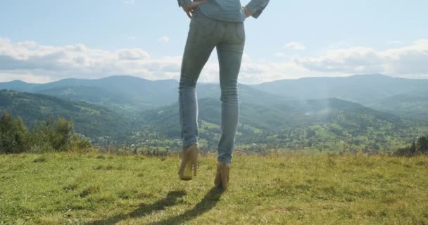 一个穿着牛仔裤和高跟鞋的女人的腿，可以看到令人叹为观止的高山和山谷。蓝天，白云，中空，白天，春天，夏天 — 图库视频影像
