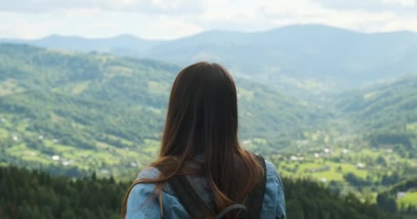 Long-haired beautiful girl turns her head and looks at the green valley and mountains with clouds. Traveler with a backpack looking at a beautiful view. Close-up, spring, summer. — Stockvideo