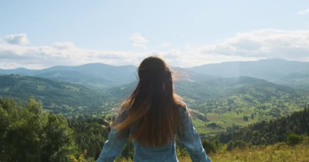 Femme se tient devant une vue imprenable sur les montagnes et la vallée verdoyante. Levant les mains dans différentes directions, le vent souffle les cheveux longs. Plan moyen, vue de dos, printemps, été, ralenti — Video