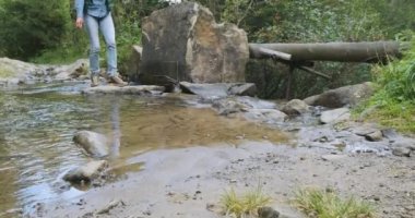 female tourist crosses a small mountain river, stops to wash his face with cool water to refresh himself. Then he continues his journey with a backpack. Spring, summer, wide shot, slow motion