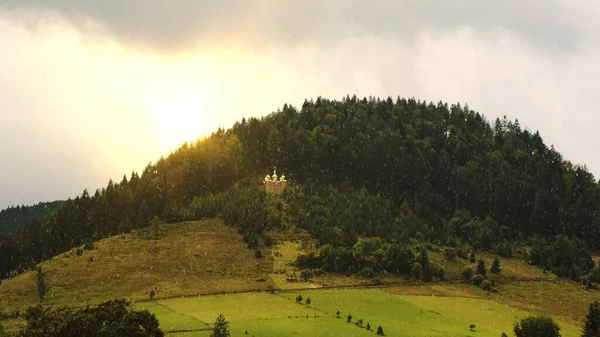 Church among the green forest on the hill. Orange sunset sun and snow — Zdjęcie stockowe