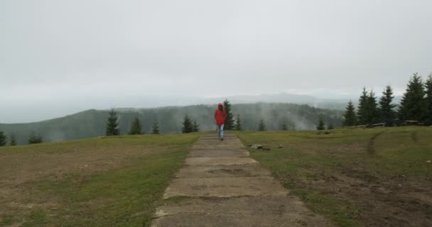 Lonely girl in a red jacket and a dressed hood, goes to meet the camera, against the backdrop of foggy mountains. Carpathians, Ukraine, Europe — стоковое видео