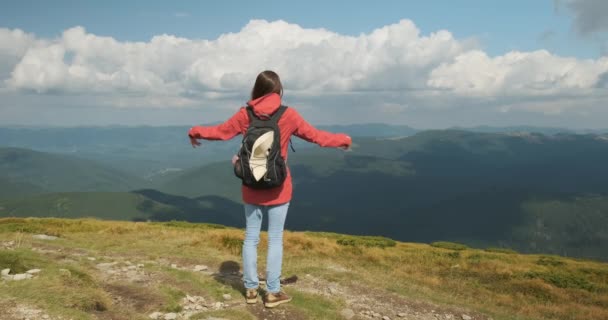 Młoda dziewczyna stoi na górze z plecakiem, rozkłada ramiona na boki i cieszy się widokiem na góry pokryte zielenią i białymi chmurami. Karpaty, Ukraina, Europa. — Wideo stockowe