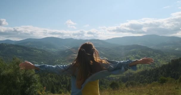 丘の上に立つ女は手を上げて風を捉える。風に髪とデニムシャツの打撃。後ろからの眺め、緑の山々と白の美しい景色 — ストック動画