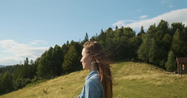 Side view of a young girl who looks into the distance while standing on a green hill. Forest in the background. Windy, blue skies, medium shot. Carpathians, Ukraine. — стокове відео