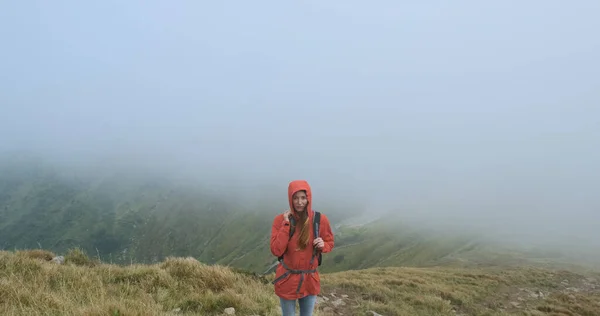 Woman look into the camera, standing on the edge of the mountain in a gray cloud. Movement of fog on the background. Stormy weather at the summit. Medium shot, spring summer, Carpathians, Ukraine. — Stockfoto