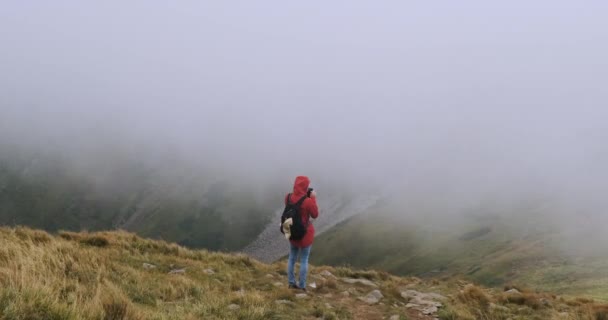 Jeune fille avec un sac à dos, se tient dans les montagnes sur le bord d'une falaise et faire quelque chose sur un smartphone, achats en ligne, correspondance. Couverture nuageuse forte, vent, mauvais temps. Carpates — Video