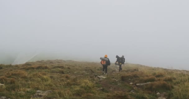 A férfi és a lány turisták, nagy, nehéz hátizsákkal, leereszkednek egy hegyről, sűrű köd borítja. Rossz idő, felhő. — Stock videók