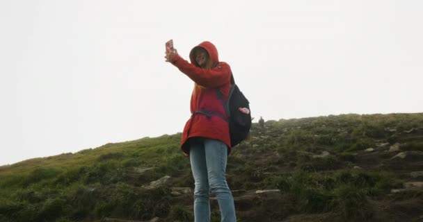 Backpacker κορίτσι στέκεται σε ένα πράσινο λόφο παίρνει μια ιστορία στο τηλέφωνό της, κοιτάζει την οθόνη και λέει γεια. Επικοινωνία στα βουνά, online internet, φωτεινός ήλιος στο φόντο του βουνού. — Αρχείο Βίντεο