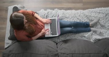 Kadın elinde dizüstü bilgisayarla kanepede yatıyor ve çalışıyor. Üst görünüm, tuşlara göre yazmak, internet sörfü. Çalışmak, evden çalışmak, serbest çalışmak..