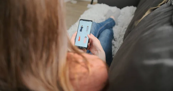 Beim Online-Shopping liegt eine Frau auf einer Couch, hält ein Smartphone in der Hand und sucht sich ein Kleid aus, während sie über den Bildschirm scrollt. Ich-Perspektive, Nahaufnahme — Stockfoto